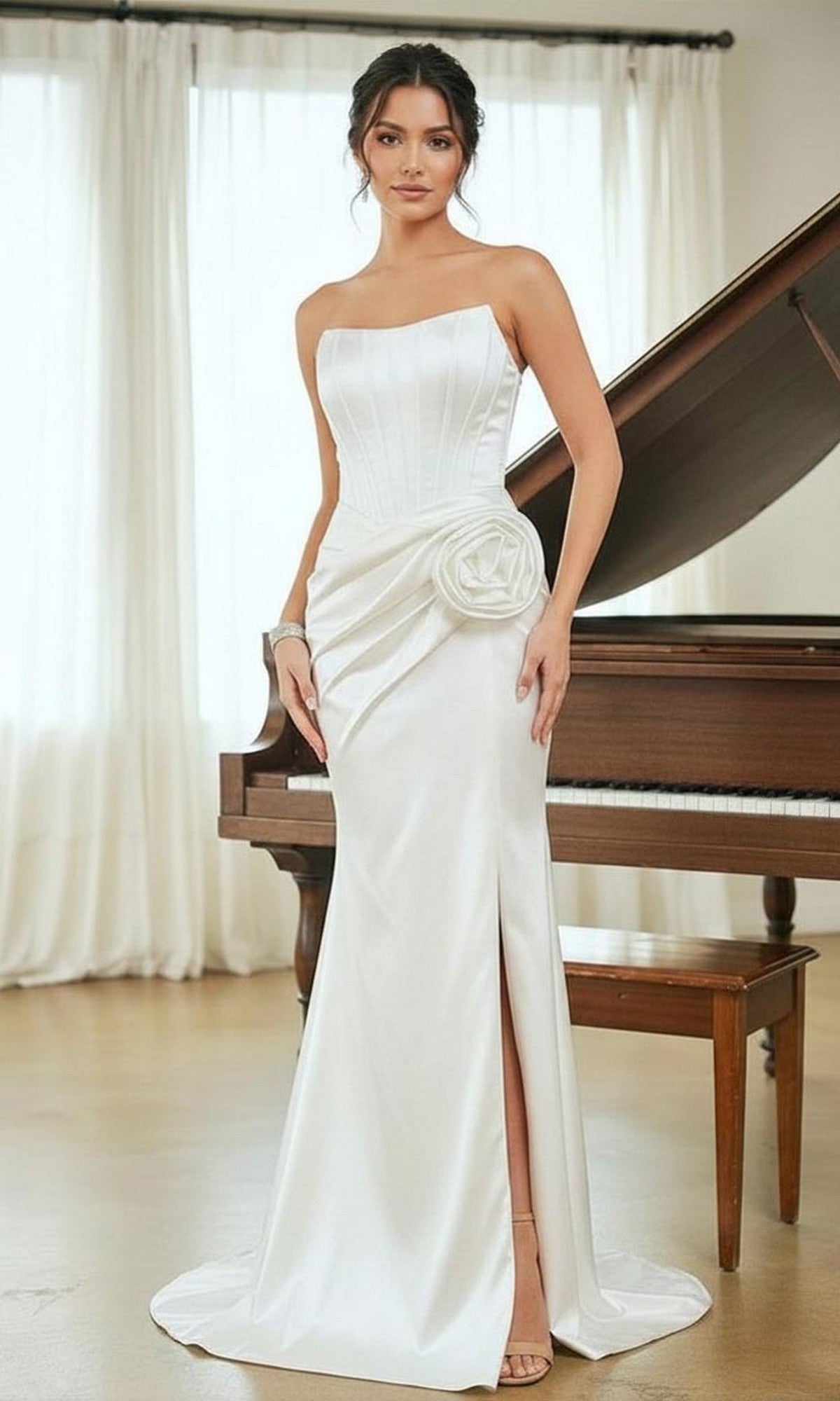 A woman stands in front of a piano wearing the Amelia Couture AL1400 long white prom dress, featuring a strapless neckline, 3D rosette at the waist, and a thigh-high slit.